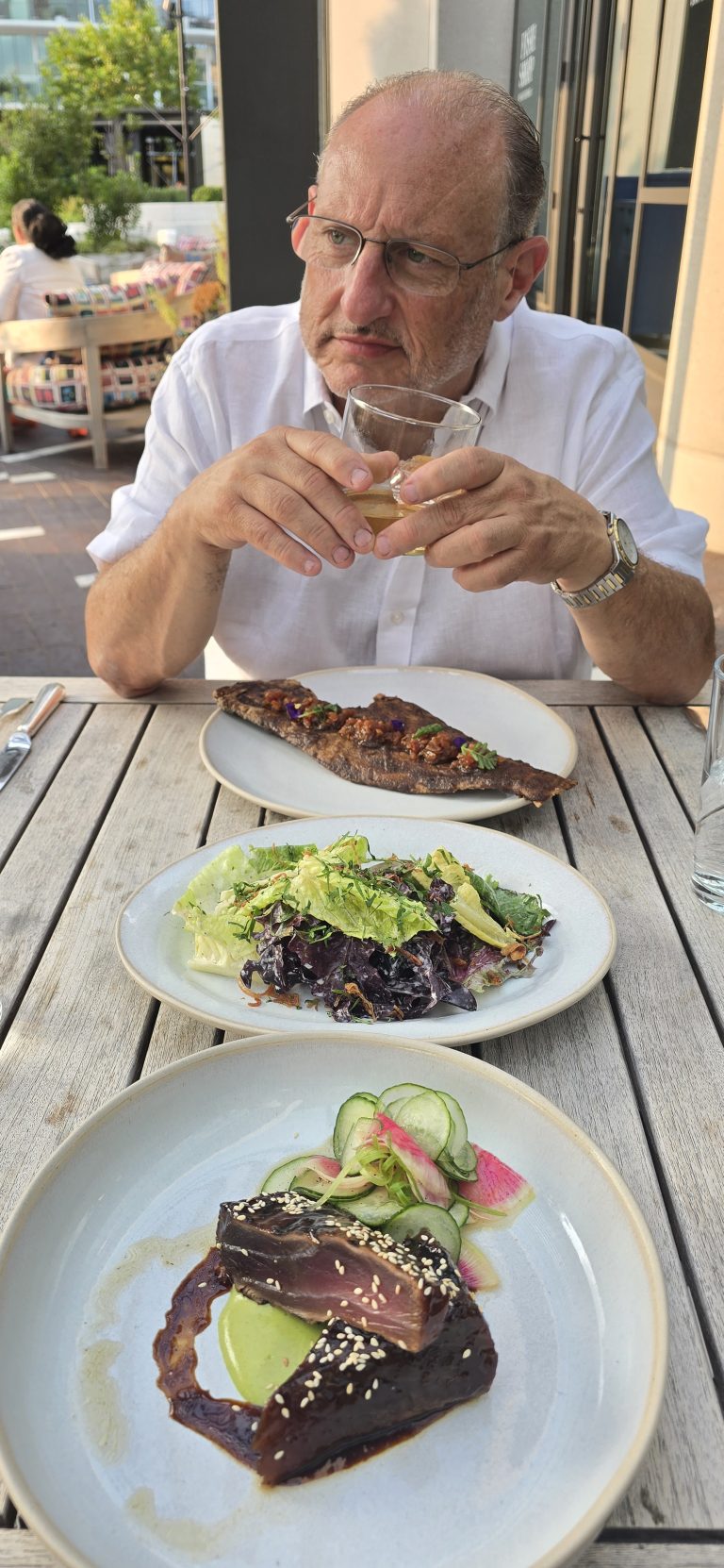 A man at a sunlit outdoor table with a glass in hand and plates of food in front of him