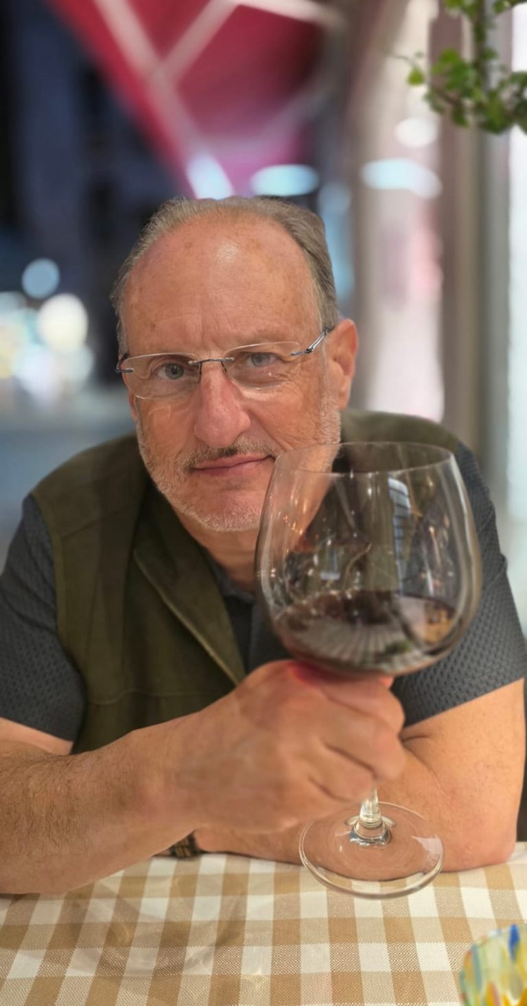 A smiling man raising an oversized glass of red wine at a checkered-cloth table